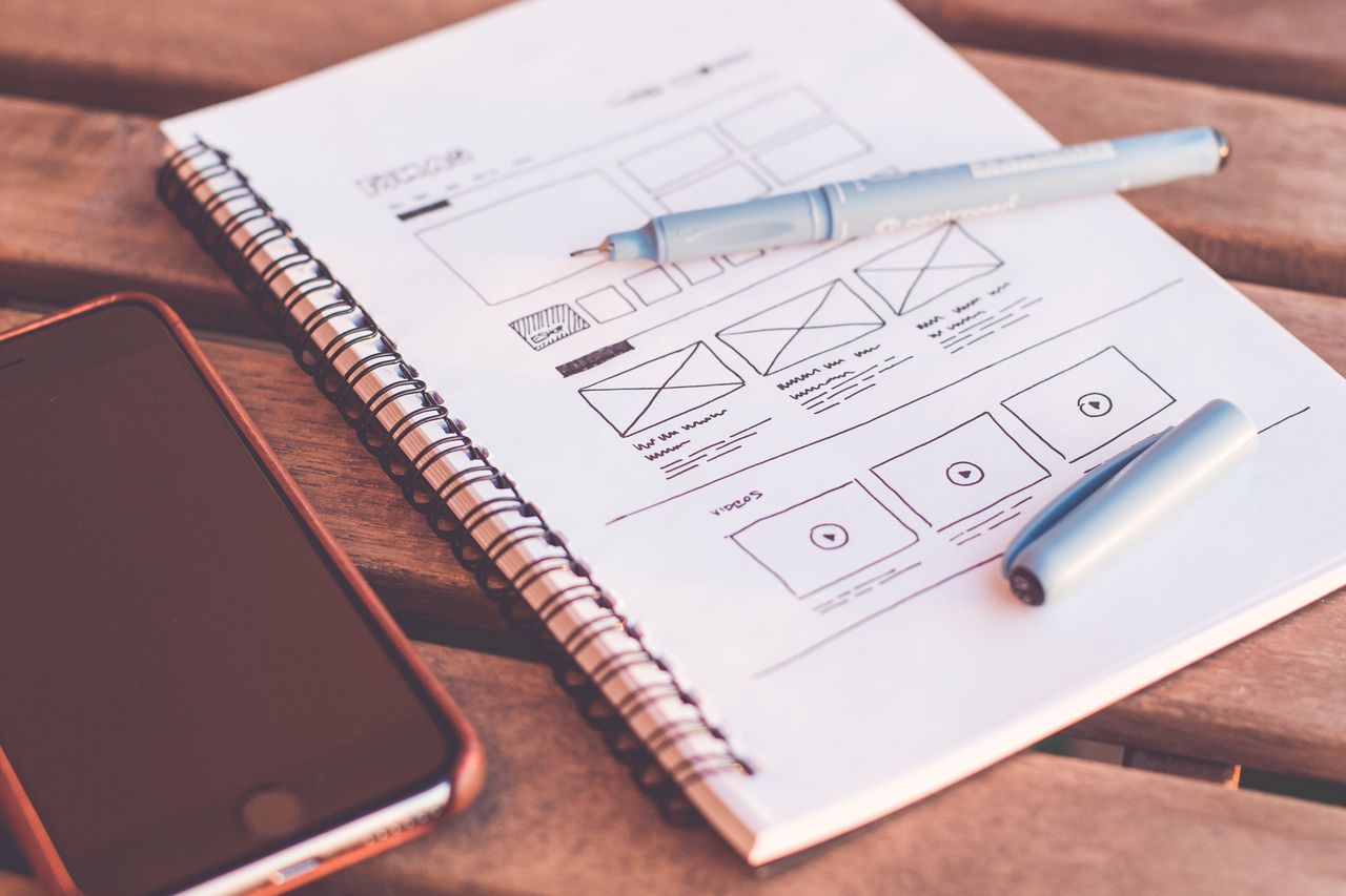 Gros plan sur un carnet avec des croquis de wireframe et un smartphone posé sur un bureau en bois.